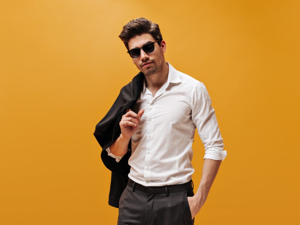 attractive-brunet-man-stylish-white-shirt-trendy-sunglasses-poses-orange-background-holds-black-jacket